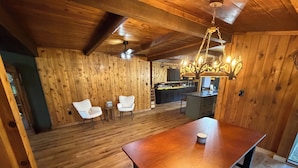 Un interior de madera con una mesa de comedor, dos sillas blancas, una araña y una zona de cocina.