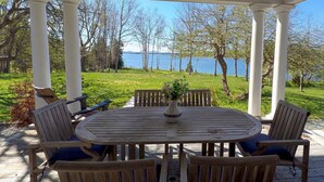 Um alpendre coberto com uma mesa de madeira redonda e cadeiras, um vaso com flores e vista para um lago e árvores.