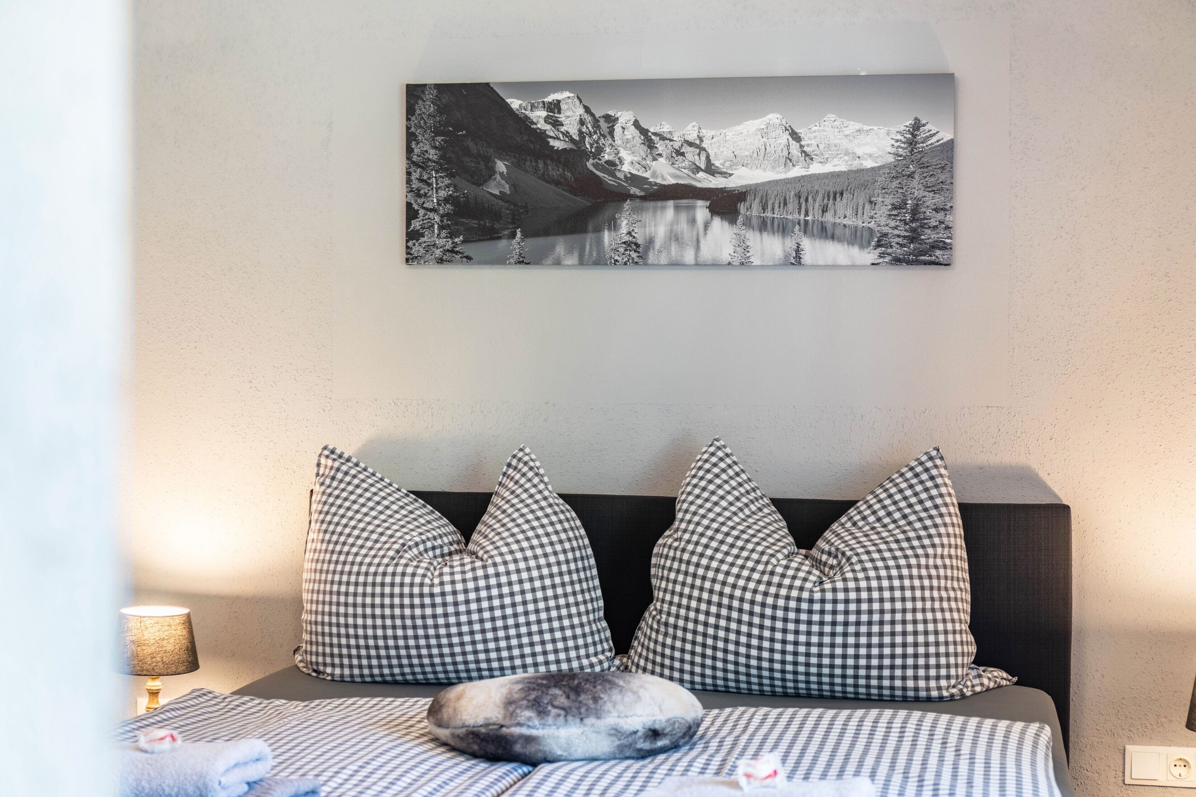 A bedroom with a bed, checkered pillows, a mountain landscape painting, and a lamp.