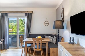 A room with a dining table, chairs, a sofa, a TV, and a view of a green landscape through a glass door. - Apartment 'Kuhglocken' with Mountain View, Shared Garden and Wi-Fi (Döbriach)