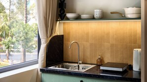 Una cocina con un lavamanos, una ventana con cortina y una estantería con cuencos y una olla.