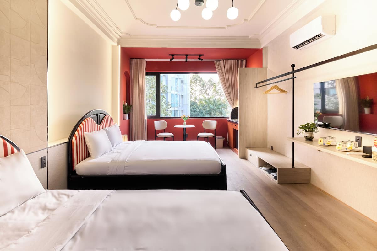 Ein Hotelzimmer mit zwei Betten, einem kleinen Tisch, Stühlen, einem Spiegel und einem Fenster mit Blick auf Gebäude und Bäume.