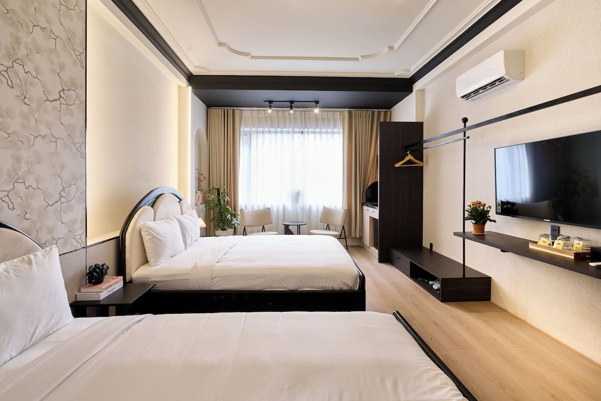 Ein Hotelzimmer mit zwei Betten, einem Fernseher, einem Schreibtisch, einem Sessel, einem kleinen Tisch und einem Fenster mit Vorhängen.