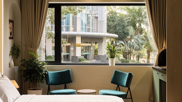 Habitación de hotel con cama, dos sillas, una mesita, ventana con vistas a edificios y árboles, y una planta en maceta.