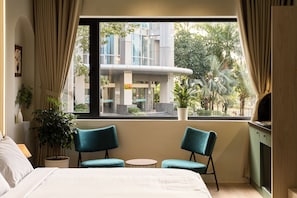 A hotel room with a bed, two chairs, a small table, a window with a view of buildings and trees, and a potted plant.