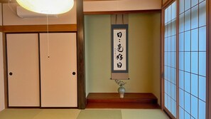 Une pièce de style japonais traditionnel, avec des portes coulissantes, un sol recouvert de tatamis, un rouleau calligraphié suspendu au mur et un vase posé sur une étagère basse en bois.