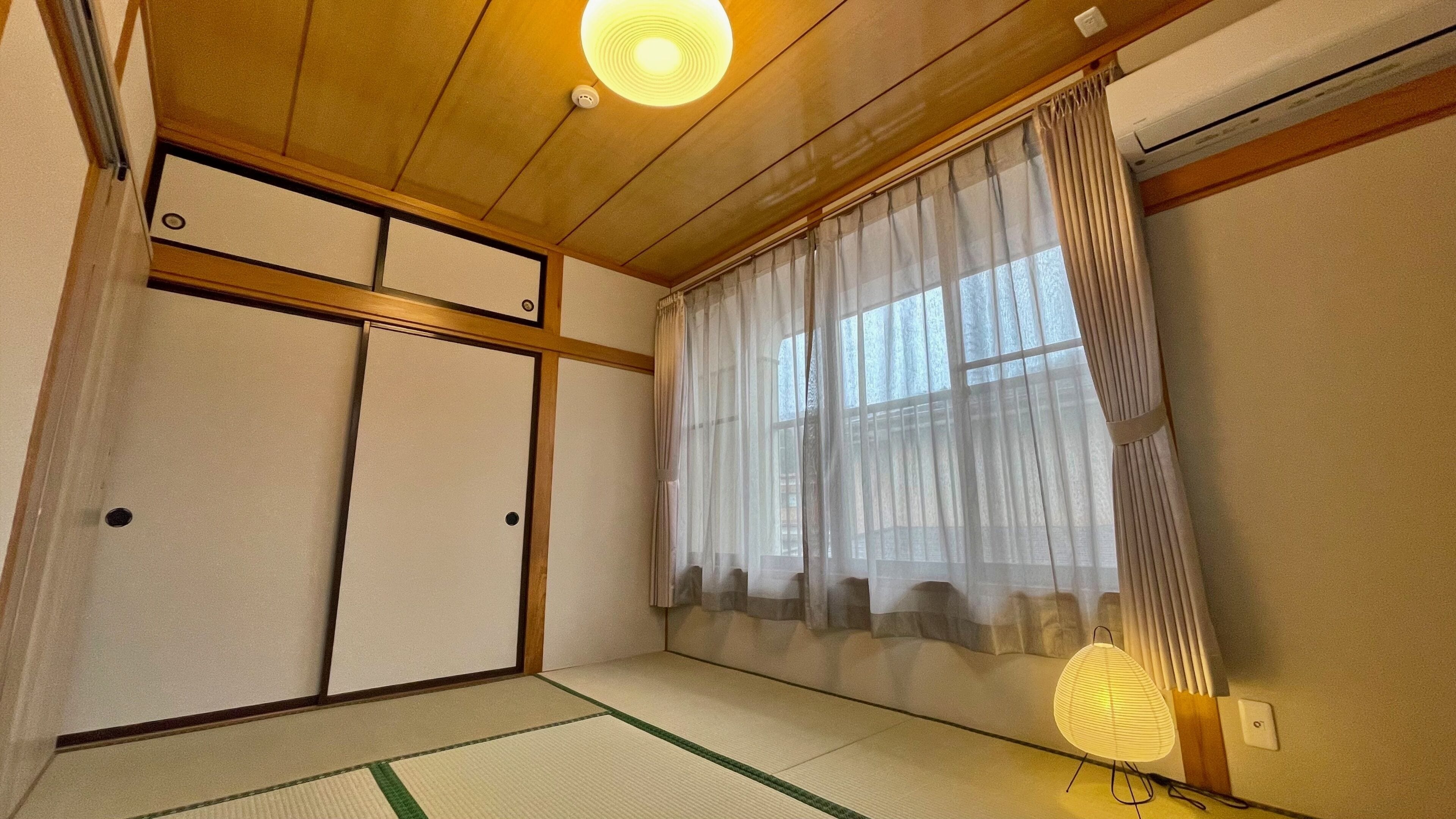 Une pièce de style japonais traditionnel, avec un sol recouvert de tatamis, des portes coulissantes, un ventilateur de plafond et une fenêtre ornée de rideaux légers.