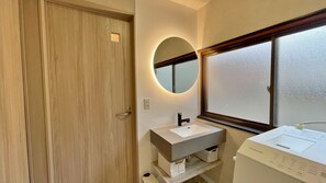 Une salle de bain avec une porte en bois, un miroir rond, un lavabo blanc et un lave-linge blanc.