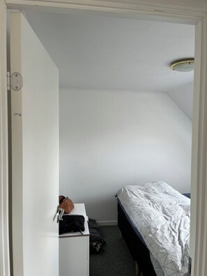 Ein kleiner, schlicht eingerichteter Raum mit einem Bett, einem Schreibtisch und einer Tür zu einem anderen Raum.