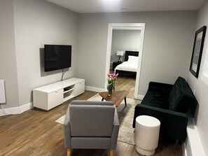 A modern living room with a flat-screen TV, a grey sofa, a coffee table with a vase of flowers, and a green armchair.