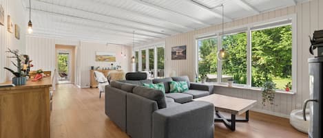 Amplia sala de estar con sofá seccional gris, piso de madera y grandes ventanas con vistas a zonas verdes.