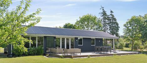 Een modern huis met een overdekte patio, een gazon en bomen op de achtergrond.