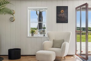 A room with a large window, a chair, a small table, a potted plant, and a framed picture on the wall.
