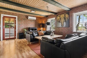 A living room with a brick wall, wooden ceiling beams, a black leather sofa, a wooden coffee table, and a large elephant painting.