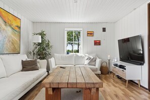 A living room with a white sofa, a wooden coffee table, a TV on a white stand, and a window with a view of trees.