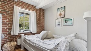 A bedroom with a brick wall, a window with curtains, a bed with floral bedding, and wall art. - Lovely holiday home in Nykøbing Mors (Nykøbing M)
