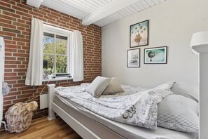 Une chambre avec un mur en briques, une fenêtre avec des rideaux, un lit avec une literie à motifs floraux et des œuvres d’art accrochées au mur.