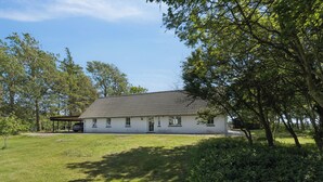 A single-story building with a sloped roof, surrounded by a grassy yard and trees. - Lovely holiday home in Nykøbing Mors (Nykøbing M)