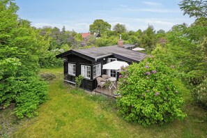 A black wooden house with white trim, surrounded by lush greenery and flowering bushes. - Cozy holiday home close to the sea in Hundested (Hundested)