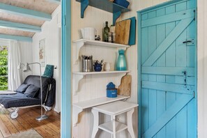Une pièce comprenant un lit, une petite table, des étagères avec des ustensiles de cuisine, une porte bleue et une fenêtre avec des rideaux.