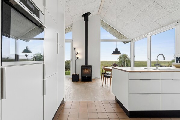 A modern kitchen with a central wood stove, large windows, and a view of the outdoors. - Cozy low-energy home with sea views (Ørsted)