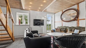 A room with a wooden ceiling, a TV mounted on the wall, a sofa with a fur throw, a wicker chair, and a round mirror. - Large charming house for 12 guests (Snøde)