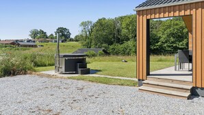 Un pavillon en bois, accessible par un chemin de gravier, entouré de verdure et surplombé d’un ciel dégagé.