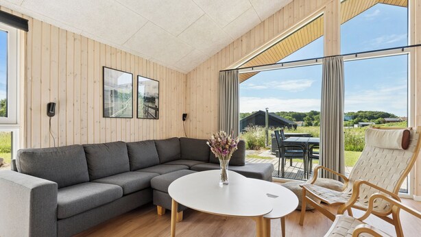 Un salon moderne avec un canapé d’angle gris, une table ronde blanche et un fauteuil en osier.