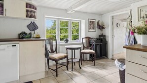 A kitchen with a dishwasher, two chairs, a small table, and a window with a view of greenery. - Idyllic Old Cottage, with low ceilings (Bredebro)