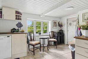 A kitchen with a dishwasher, two chairs, a small table, and a window with a view of greenery. - Idyllic Old Cottage, with low ceilings (Bredebro)