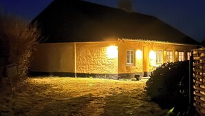 A house with a stone facade, a dark roof, and a well-lit front porch. - Idyllic Old Cottage, with low ceilings (Bredebro)