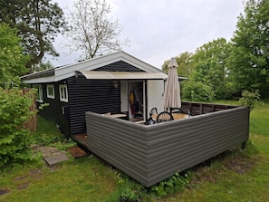 Eine kleine, eingeschossige Holzhütte mit einer Terrassenfläche, einem überdachten Sitzbereich im Freien und einem sichtbaren Kleiderständer.