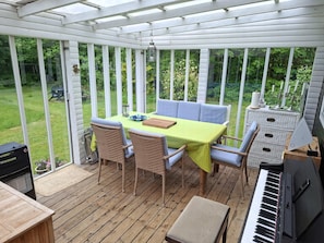 Eine überdachte Terrasse mit Tisch, Stühlen, einem Klavier und Blick auf einen grünen Garten.