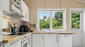 Moderni keittiö, jossa on valkoiset kaapit, puinen työtaso, sisäänrakennettu astianpesukone ja ikkuna, josta avautuu näkymä vehreään maisemaan.