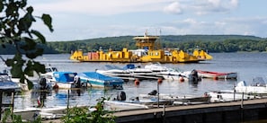 A ferry dock with multiple boats, a large yellow ferry, and a forested hill in the background. - Cottage on the island IVÖ in Skåne (Fjälkinge)