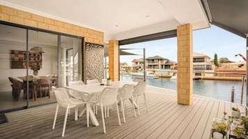 A modern outdoor dining area with a wooden deck, a glass partition, and a view of the water and buildings.