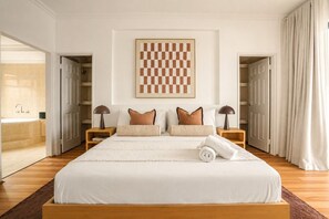 Ein Hotelzimmer mit einem großen Bett, zwei Nachttischen, einem gerahmten Kunstwerk über dem Bett sowie Blick auf ein Badezimmer und einen Flur.