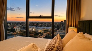 Hotelkamer met een groot raam met uitzicht op de stad bij zonsondergang, een bed met kussens en een lamp.
