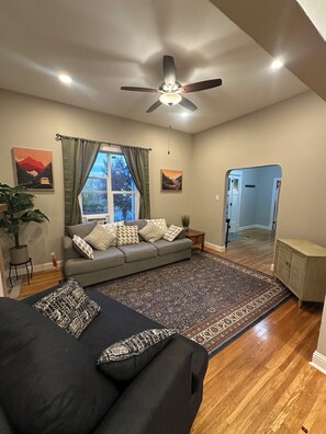 Ein Wohnzimmer mit Sofa, Teppich, Deckenventilator und einem Fenster mit Vorhängen.