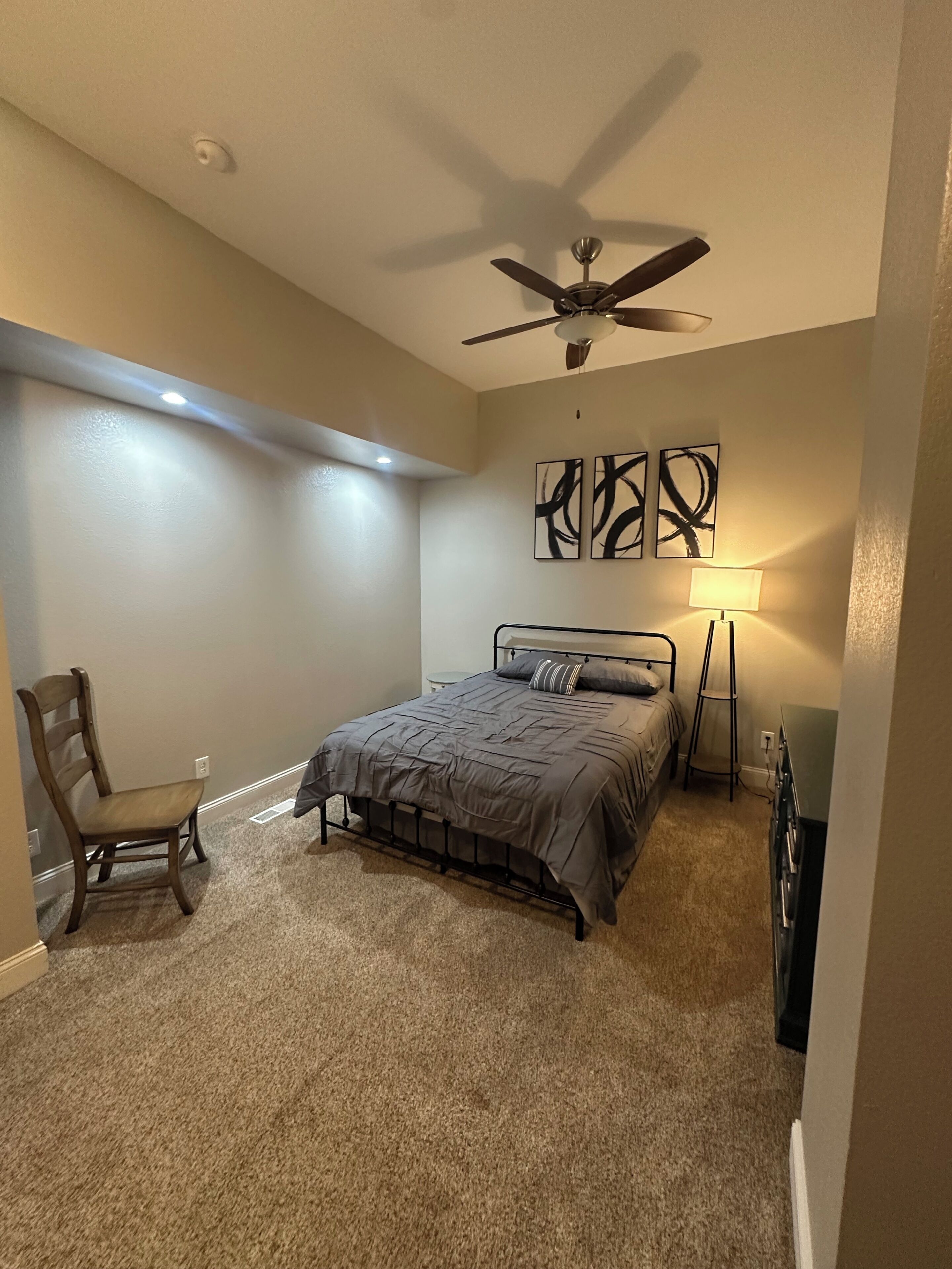 A bedroom with a bed, a chair, a ceiling fan, and abstract wall art.
