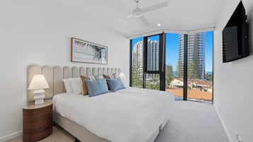 Quarto com cama, luminárias de cabeceira, ventoinha no teto, uma janela grande com vista para a cidade e TV na parede.
