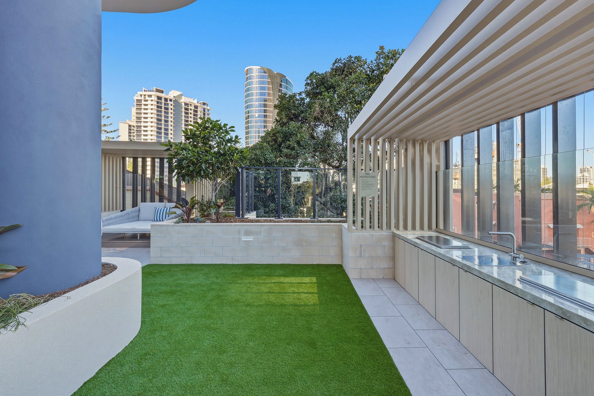 A modern outdoor area with a green artificial turf, a seating area with a sofa and cushions, and a glass-enclosed space with a view of the cityscape.