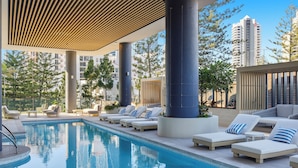 A poolside area with lounge chairs and a swimming pool, surrounded by tall buildings and trees. - Amaya (Broadbeach)
