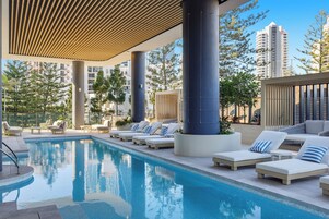 Zona de piscina con sillas de descanso y una piscina, rodeada de edificios altos y árboles.
