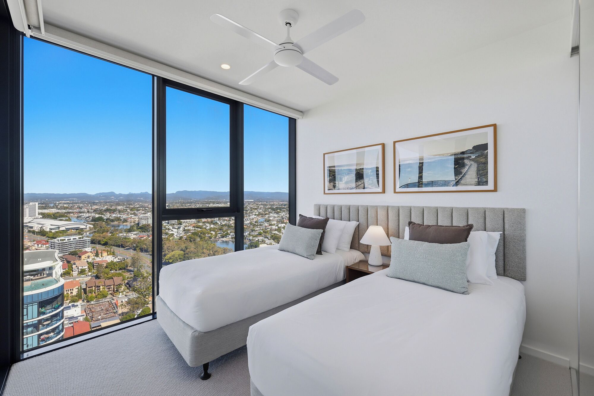 A hotel room with two beds, a ceiling fan, and large windows offering a city view.