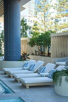 Zona de piscina con sillones de descanso, un pilar grande y vistas de edificios circundantes.