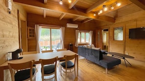 A wooden cabin interior with a dining table, chairs, a sofa, and a TV mounted on the wall.