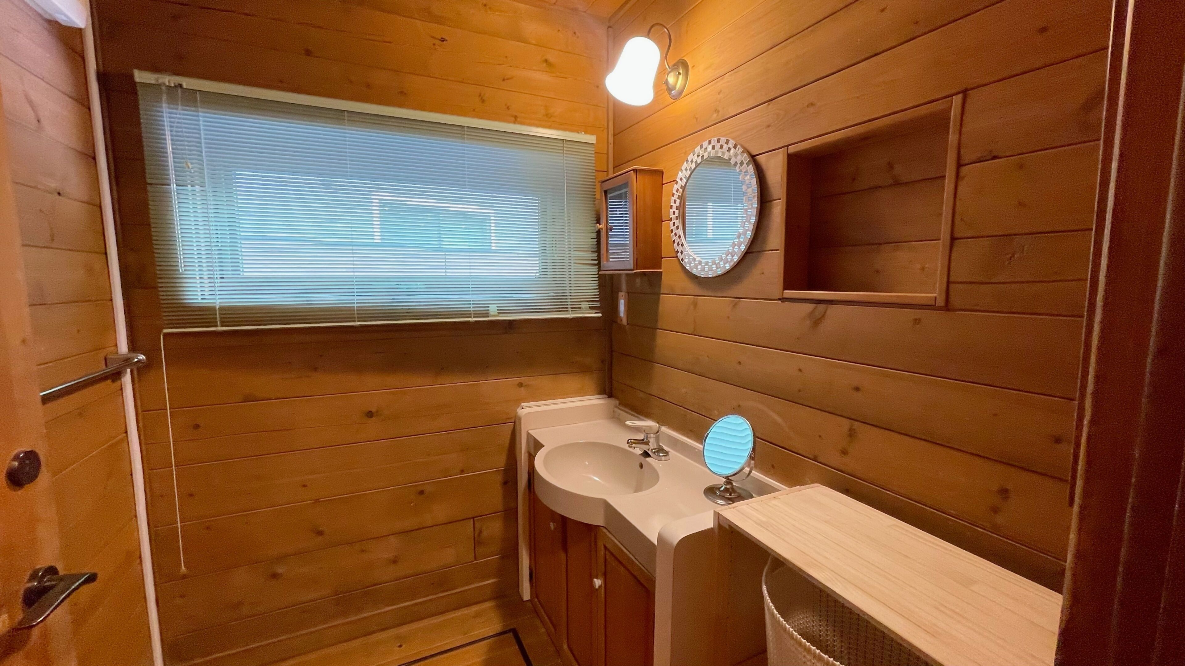 Een houten badkamer in een hutje, met een raam, een wastafel, een spiegel en een handdoekhouder.