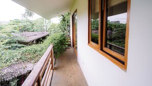 Un balcón con baranda de madera, piso de concreto y vistas a abundante vegetación y un edificio distante.