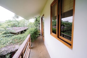 Un balcón con baranda de madera, piso de concreto y vistas a abundante vegetación y un edificio distante.